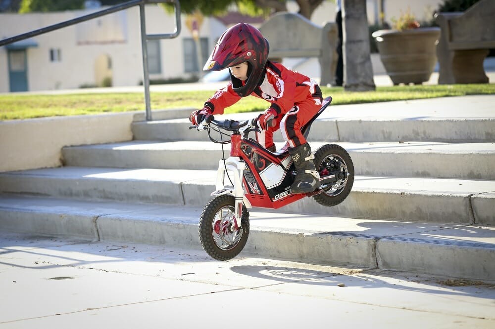4 year old girl riding a small electric motorbike down four concrete steps and wearing a full-face crash helmet, riding gear, gloves and boots.  